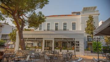An outdoor cafe area with tables and chairs, a large tree, and a building with a balcony.