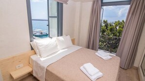 A hotel room with a bed, a small wooden bench, and a view of the sea and boats through large windows. - Panellinion Luxury Rooms (Kalamata)