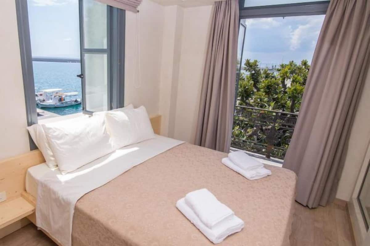 A hotel room with a bed, a window with a view of the sea and a boat, and a balcony with plants.