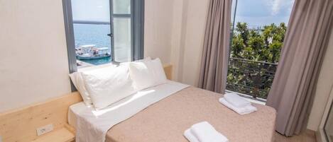 Une chambre d’hôtel avec un lit, un petit banc en bois et une vue sur la mer et les bateaux, grâce à de grandes fenêtres.