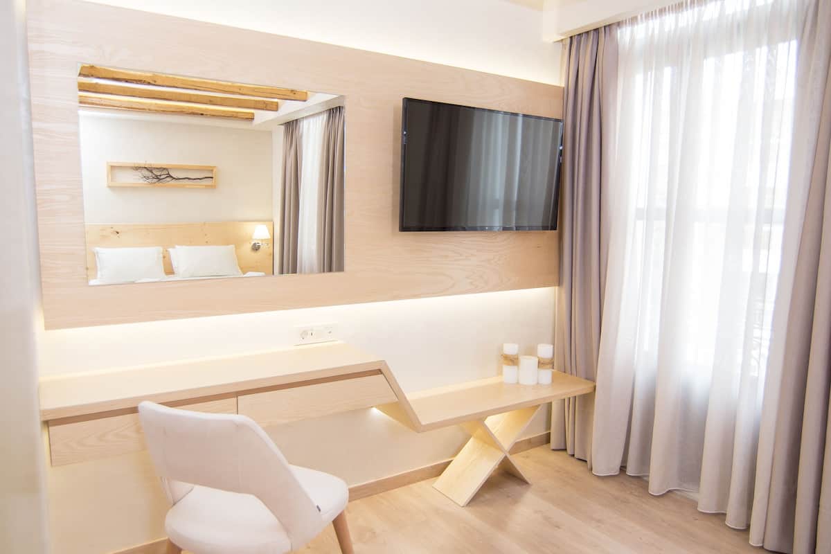 A modern bathroom with a wooden vanity, a large mirror, a flat-screen TV, and a white chair.