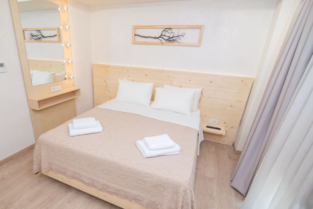 A neatly made bed with white linens and two folded towels, a wooden headboard, a bedside table with a lamp, and a framed picture on the wall.