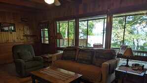 L’intérieur d’un chalet accueillant, avec un plancher en bois, un canapé, un fauteuil, une table et une fenêtre donnant sur les arbres.