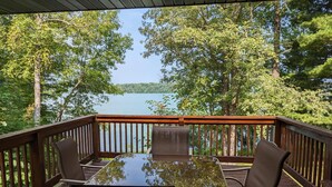 Une terrasse en bois avec une table en verre et quatre chaises, donnant sur un lac entouré d’arbres.