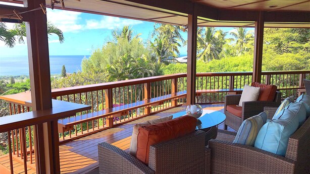 A covered outdoor seating area with wicker furniture and a wooden railing, overlooking a lush landscape and the ocean.
