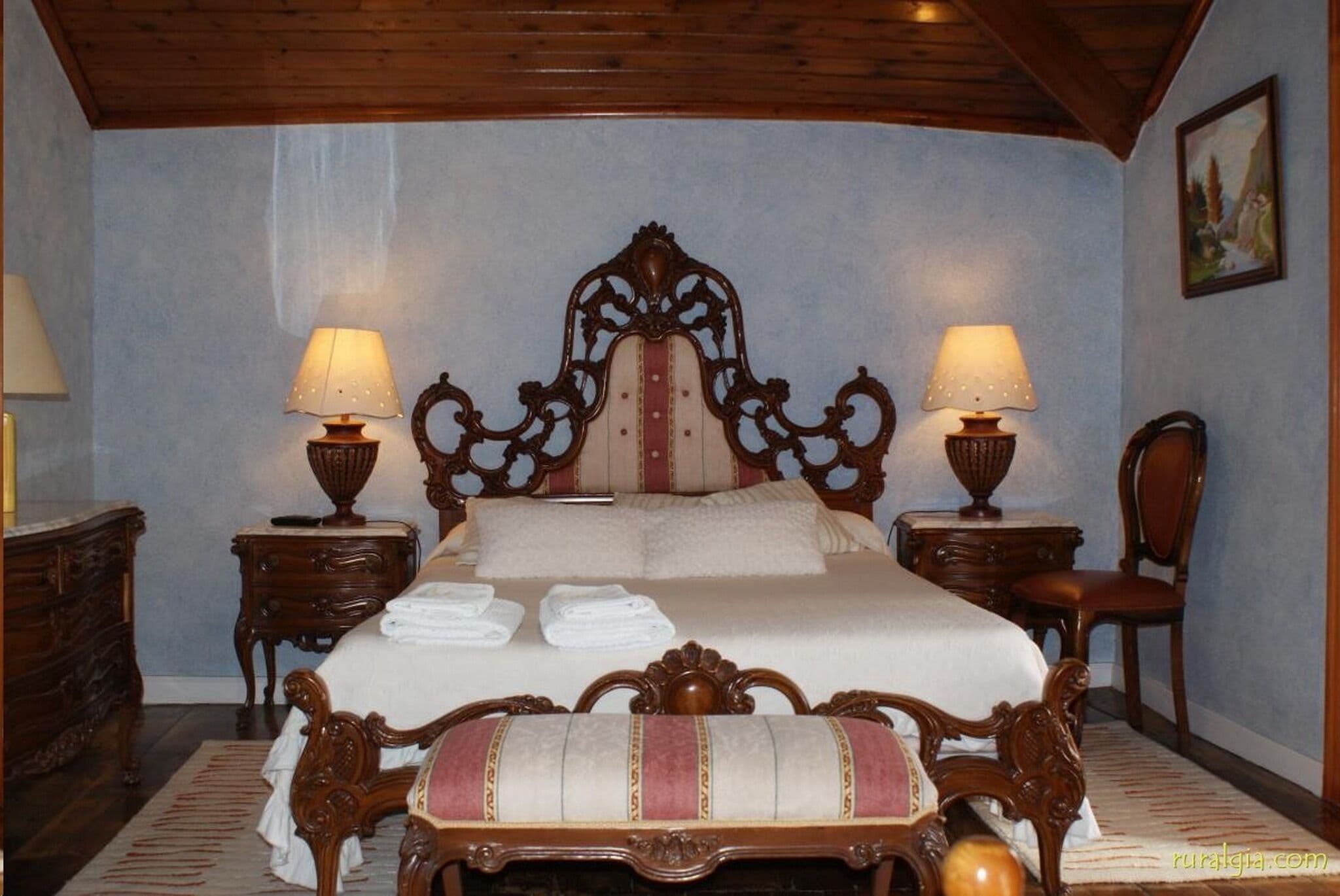 Un dormitorio tradicional con una cama grande, dos mesitas de noche, una silla de madera y un cuadro en la pared.