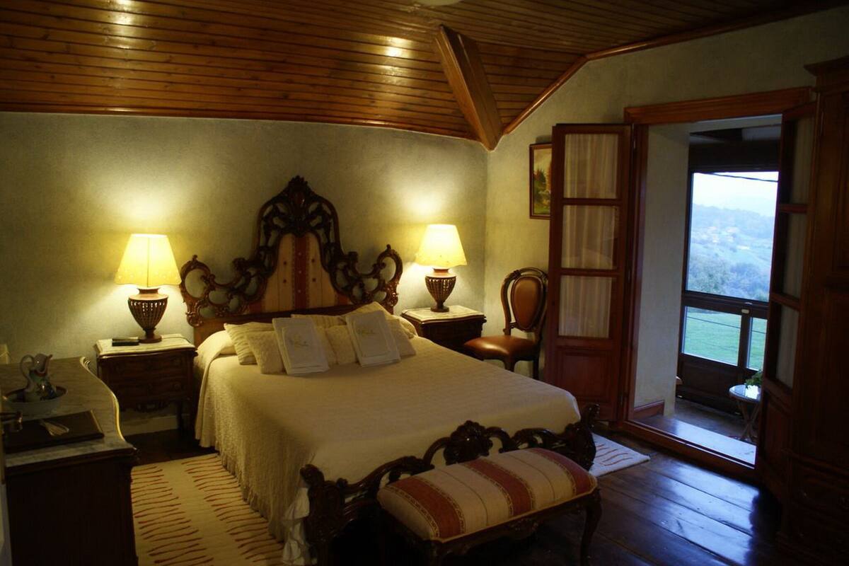 A bedroom with a wooden ceiling, a large bed with two bedside lamps, a chair, and a view through an open door.