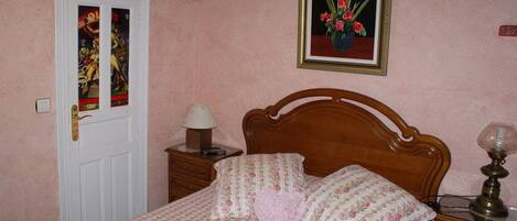 A bedroom with a wooden bed, floral bedding, a nightstand with a lamp, a framed picture on the wall, and a door with a stained glass window.