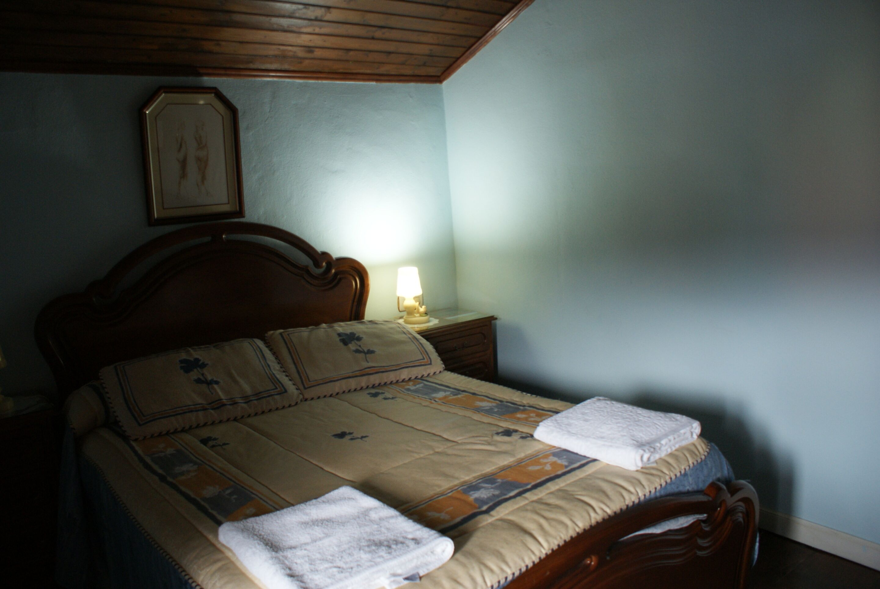 Una cama bien hecha con una colcha y dos toallas, una lámpara de noche y un cuadro en la pared.