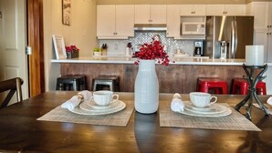 Un coin repas avec une table en bois dressée pour deux personnes, un vase blanc contenant des baies rouges, et une cuisine en arrière-plan.