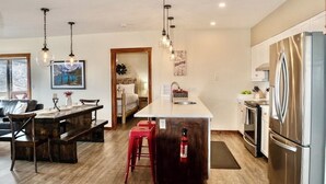 Une cuisine moderne dotée d’un îlot central, d’appareils électroménagers en acier inoxydable, de luminaires suspendus et d’un coin repas avec une table en bois et des tabourets rouges.