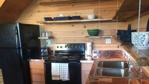 A compact kitchen with a black refrigerator, a stove, and a sink. There are wooden shelves above the stove holding dishes and a green bowl. - Rural Respite & Retreat Area (Fitzpatrick)