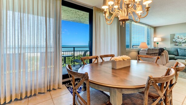 A dining area with a round table, wooden chairs, a chandelier, and a view of the ocean through large windows. - Beautiful Oceanfront, 3br/2ba Beachwalker (Amelia Island)