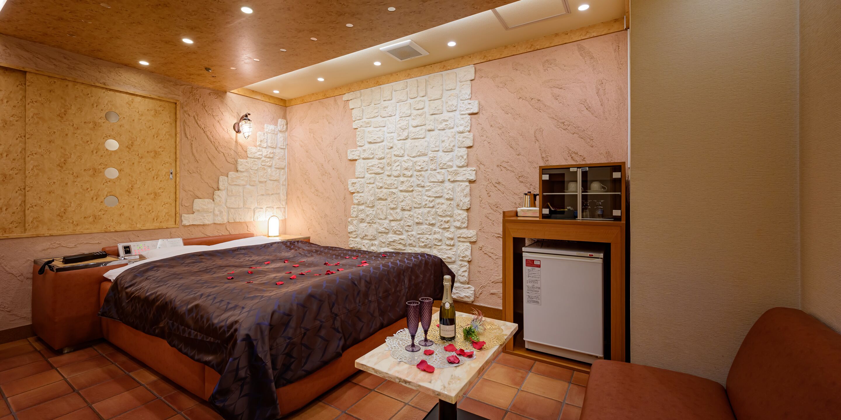 A hotel room with a bed, a small table with a bottle and glasses, a refrigerator, and a stone wall.