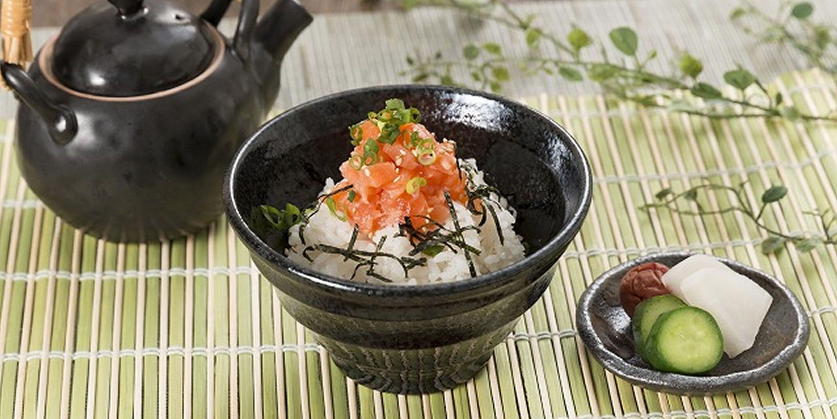 A bowl of sushi with toppings, a teapot, and a small dish with cucumber and a white item on a bamboo mat.