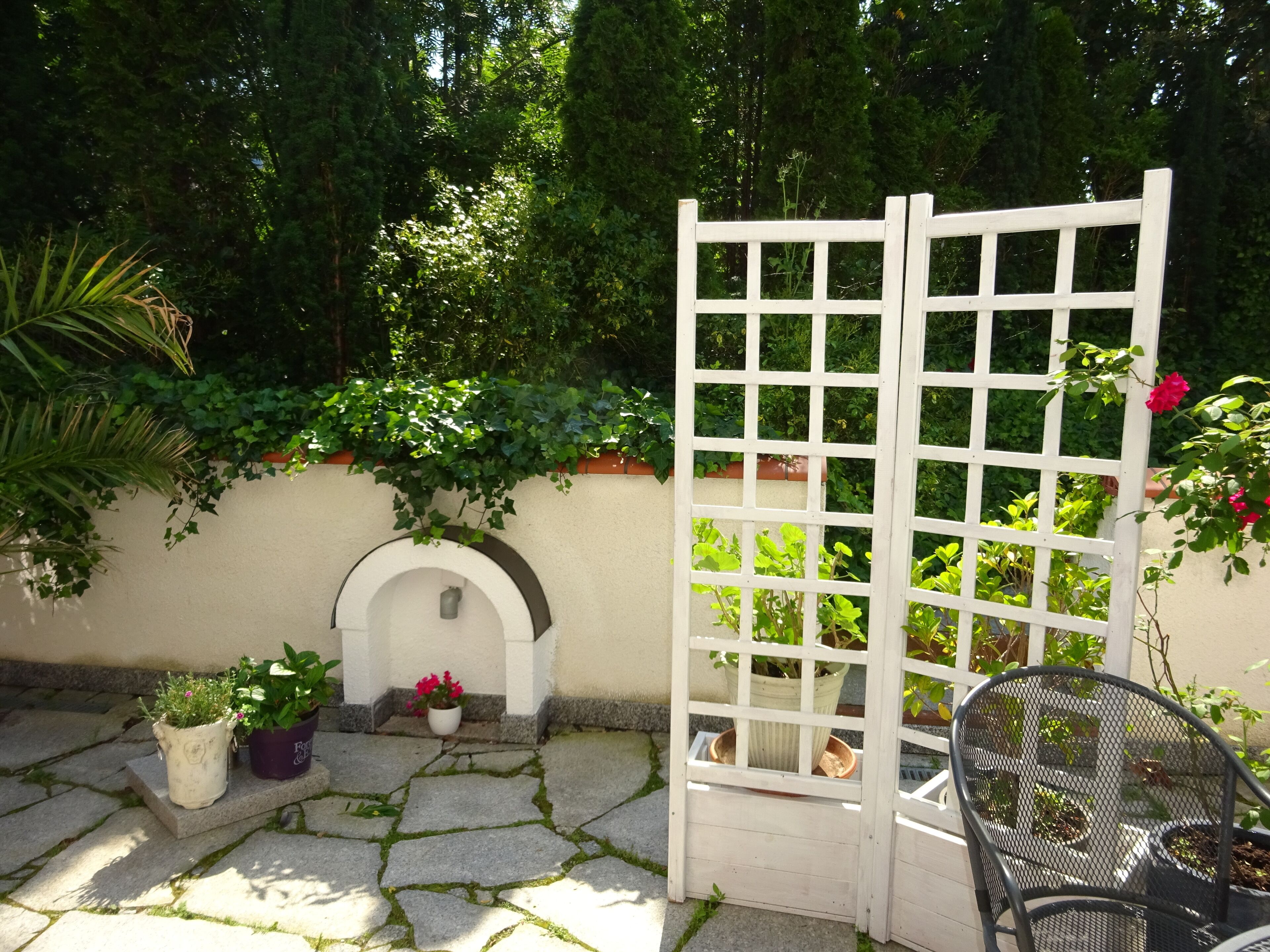 A stone-paved patio with a white trellis, potted plants, and a metal chair.