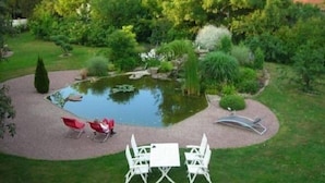 A garden with a pond, surrounded by greenery and a patio with white chairs and a table.