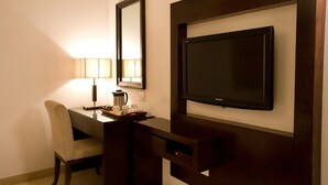 A hotel room with a dark wooden desk, a chair, a flat-screen TV mounted on the wall, and a lamp on the desk. - Eddison Hotel (Gurugram)
