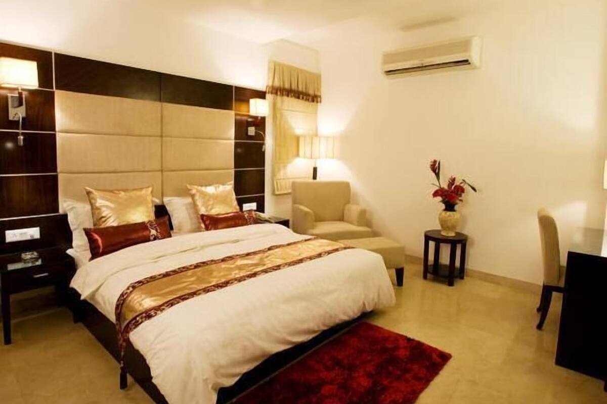 A hotel room with a large bed, a desk, a chair, an air conditioning unit, and a lamp.