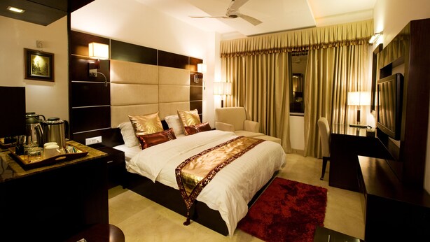 A hotel room with a large bed, bedside tables, a desk with a chair, a TV, and a ceiling fan. - Eddison Hotel (Gurugram)