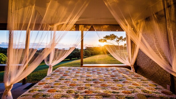 Une chambre à coucher avec un lit, des rideaux légers et une vue sur un coucher de soleil se déclinant sur un paysage.