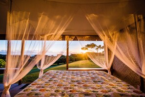 A bedroom with a bed, sheer curtains, and a view of a sunset over a landscape.