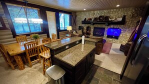 Cocina moderna con encimeras de granito, mesa y sillas de madera. Hay una chimenea de piedra, un televisor y un refrigerador.