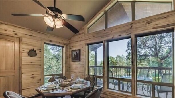 A wooden cabin interior with a dining table set for four, a ceiling fan, and large windows offering a view of the outdoors. - Tucked Away Cabin | Quiet Forest Haven w/ Hot Tub Deck (Ruidoso)
