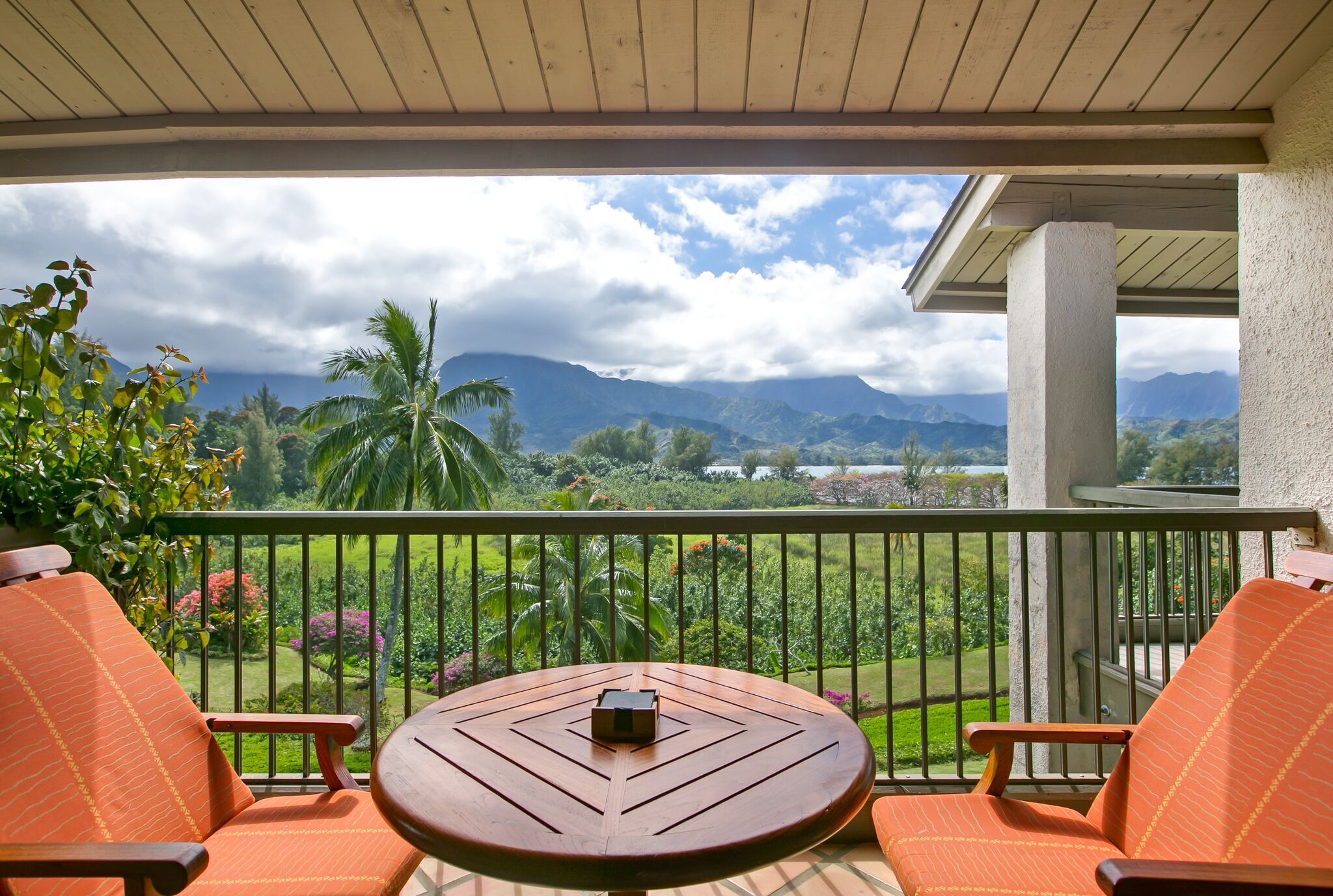 Un balcon avec une table en bois et des chaises orange, donnant sur un jardin luxuriant et des montagnes.