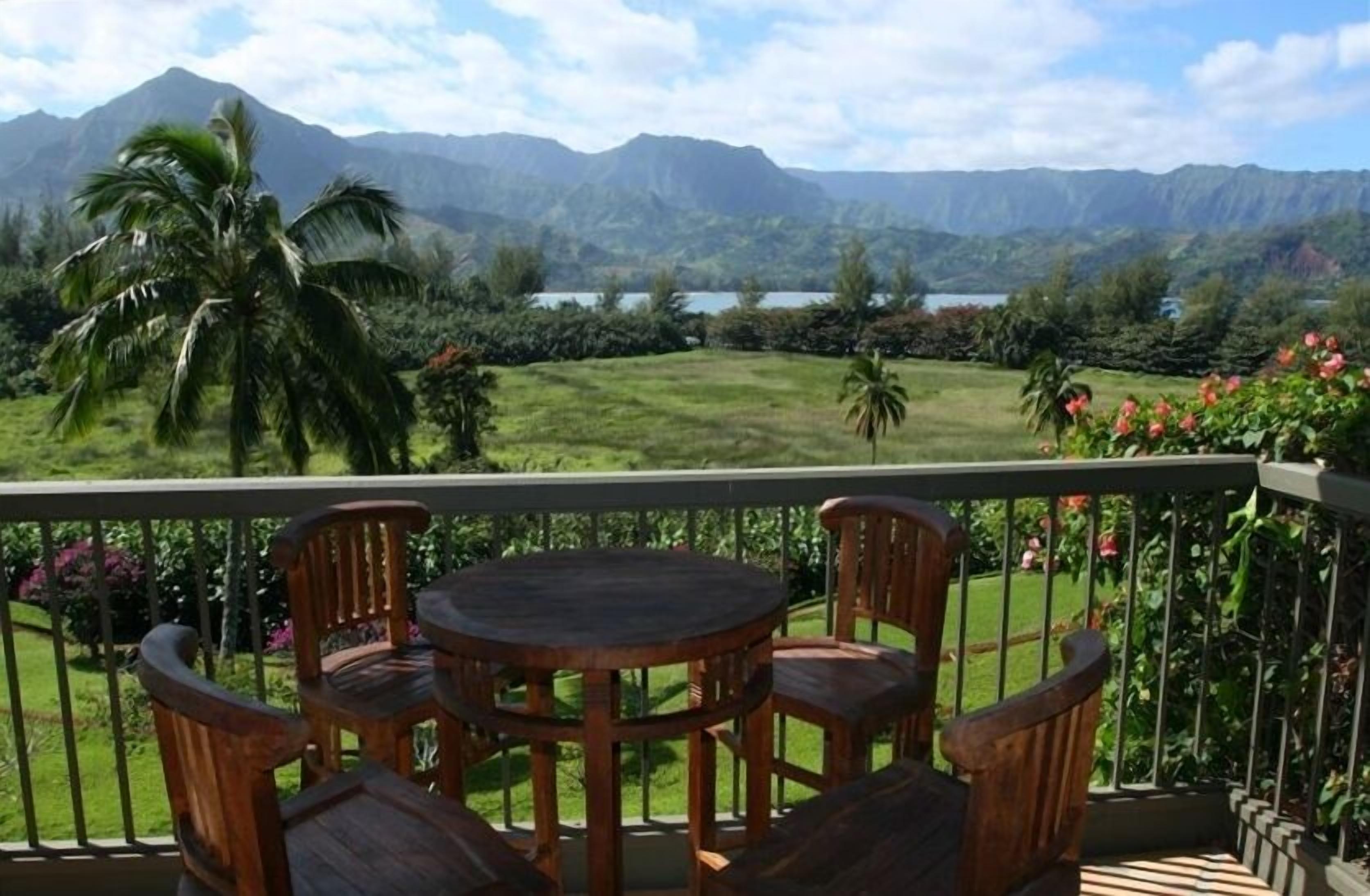 A balcony with a round wooden table and four chairs, overlooking a lush landscape with mountains and a lake.