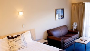 Quarto de hotel com uma cama, um sofá de couro marrom, uma mesinha com um copo e um ventilador fixado na parede.
