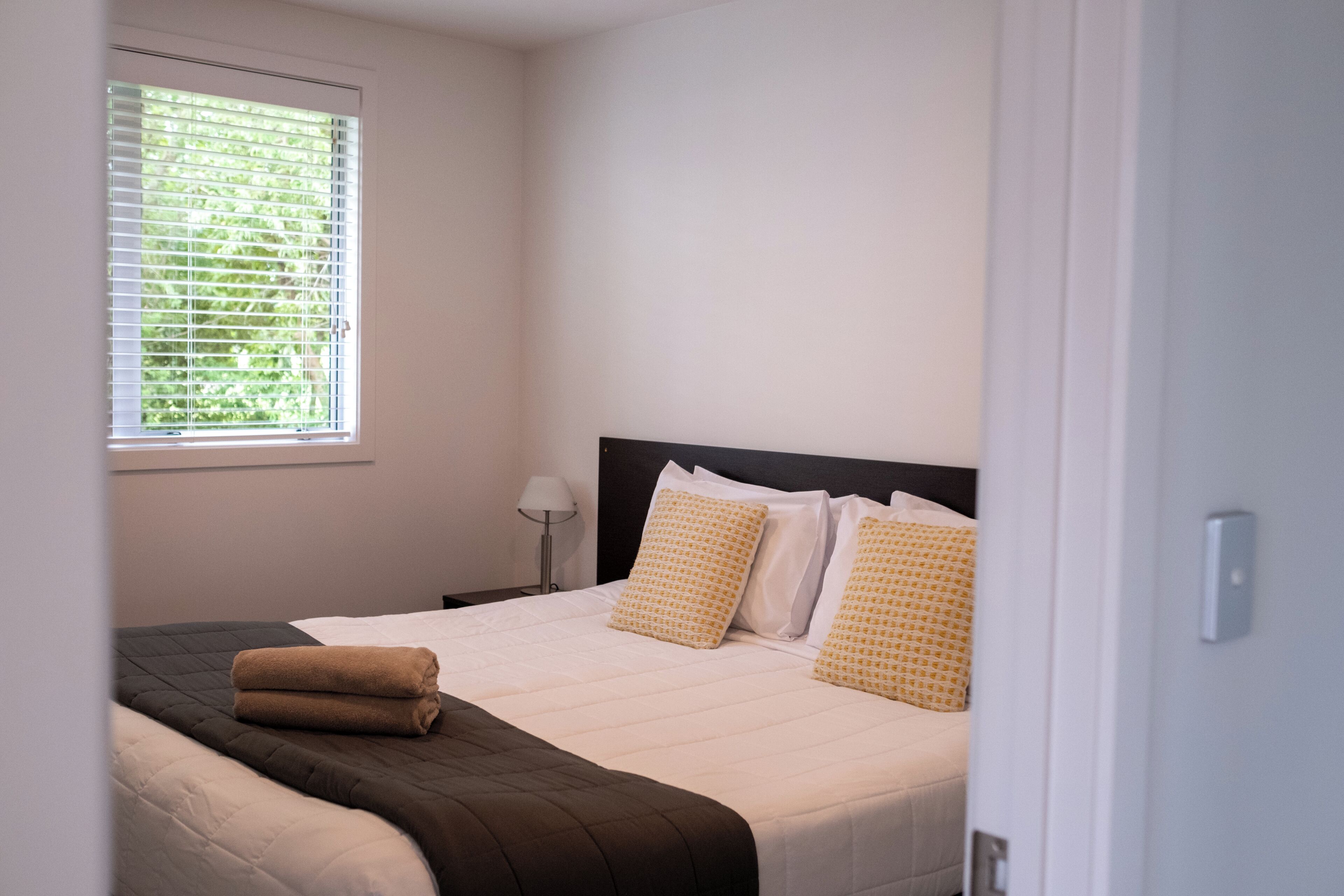 A neatly made bed with a brown blanket, a window with blinds, and a bedside lamp.