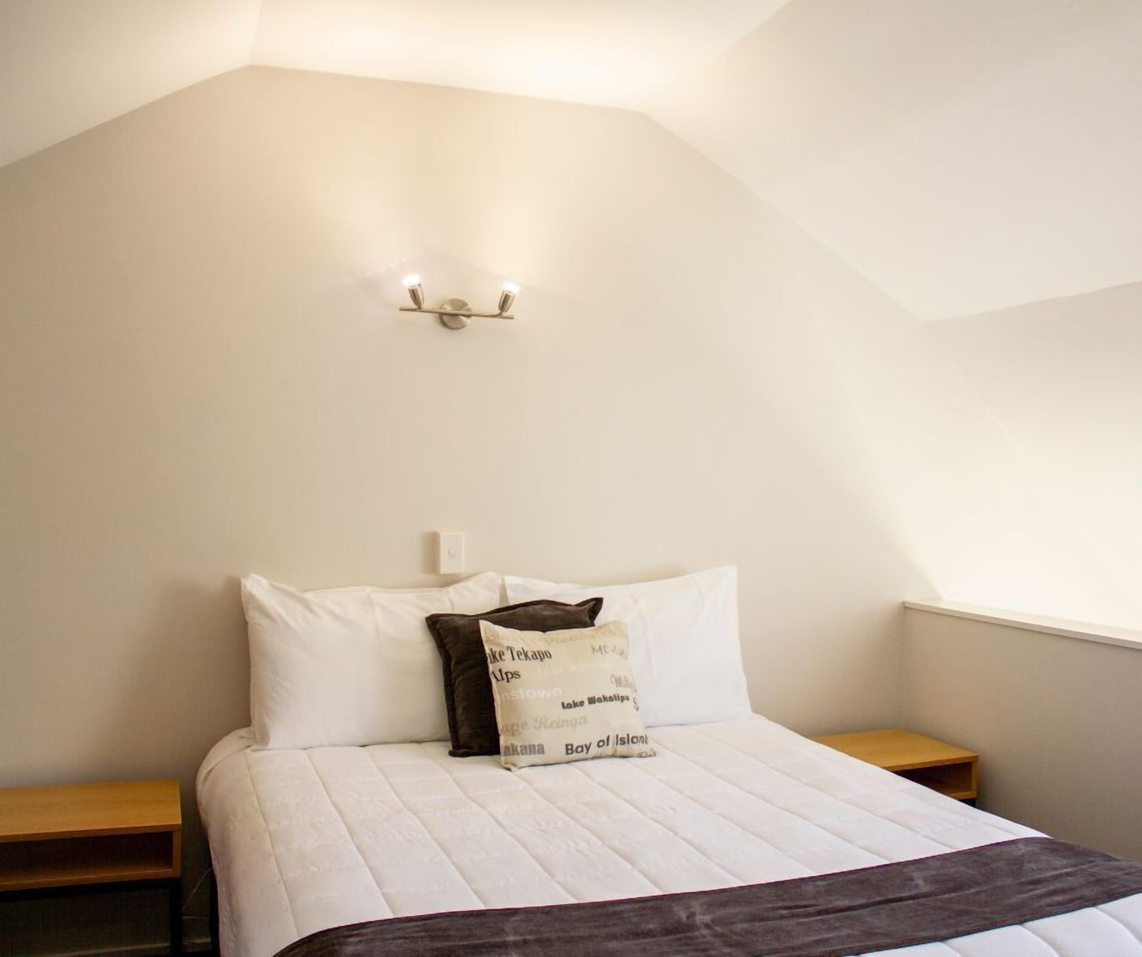 A bedroom with a bed, two bedside tables, a wall-mounted light fixture, and a window.