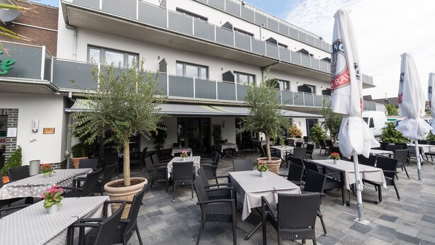 Un área de café al aire libre con mesas y sillas, un edificio con balcones y plantas en macetas.