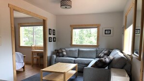 Un salón con un sofá gris, una mesa de centro de madera y un mueble blanco pequeño. Hay dos ventanas que ofrecen luz natural, y una puerta que da a otra habitación.