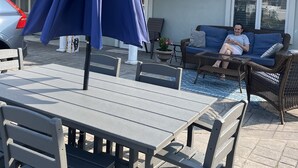 Un patio al aire libre con una mesa, sillas, un paraguas azul y una persona sentada en un sofá.