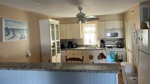 Una cocina con encimera de granito, armarios blancos, ventilador de techo, microondas, estufa y refrigerador.