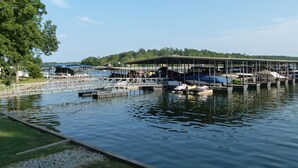 Un puerto deportivo con barcos amarrados bajo una zona cubierta, una orilla verde y árboles al fondo.