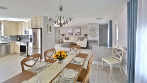 Una cocina moderna con electrodomésticos de acero inoxidable, un comedor con una mesa de madera dispuesta para cuatro personas, y una sala de estar con sofá y mesa de centro.