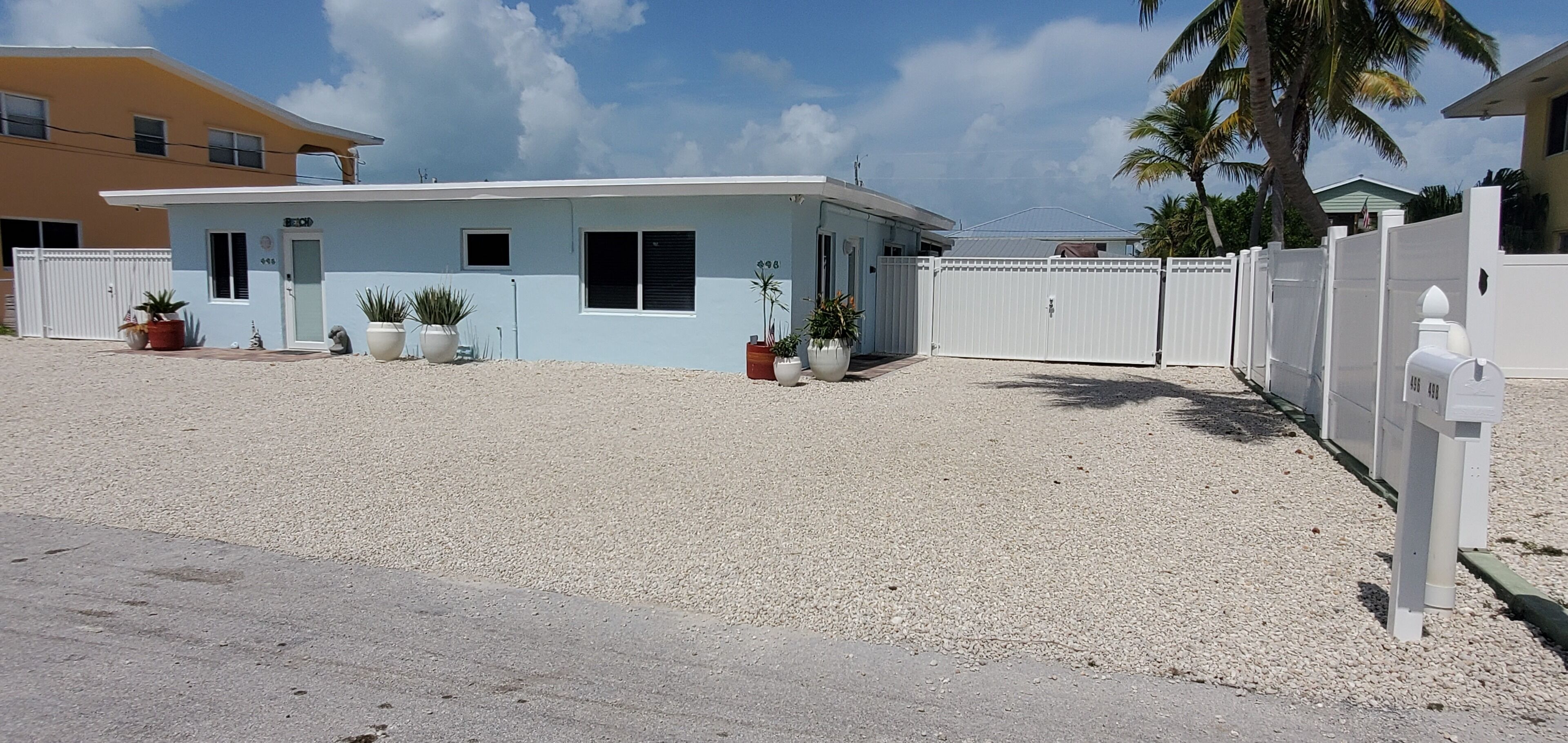 A small, light blue building with a white fence, surrounded by potted plants and a gravel driveway.