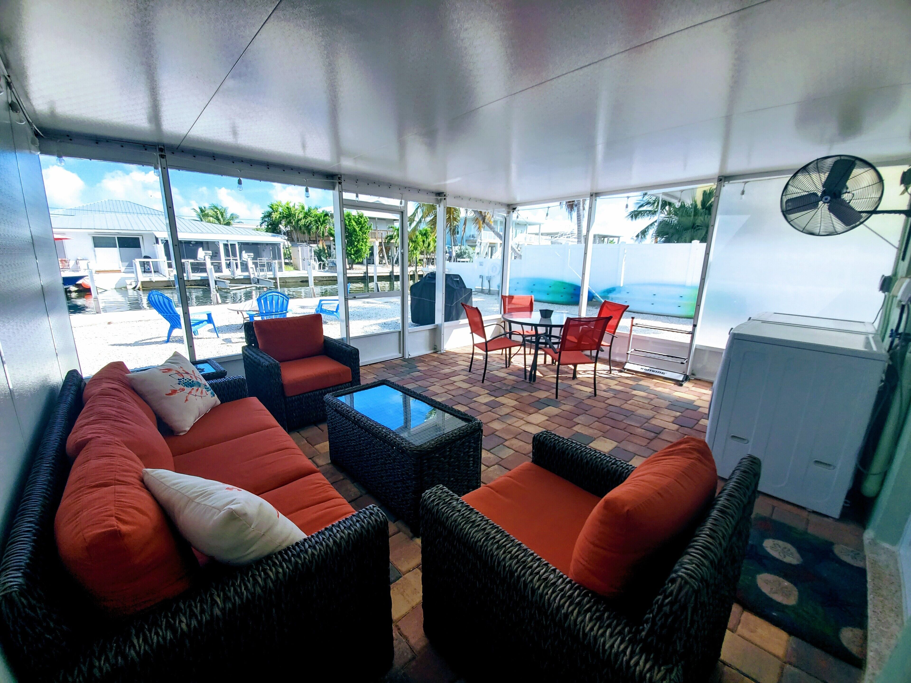 A covered outdoor seating area with wicker furniture, a fan, and a view of a pool and buildings.