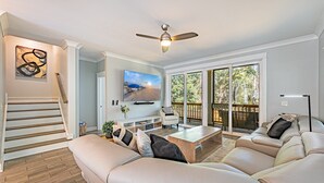 A spacious living room with a large beige sofa, a coffee table, and a television. There is a staircase leading to another level, a balcony with a view of trees, and a ceiling fan. - 117 Heated Pool | Elevator | Kids Retreat | Sleeps Big Families | Pet Friendly (Hilton Head Island)