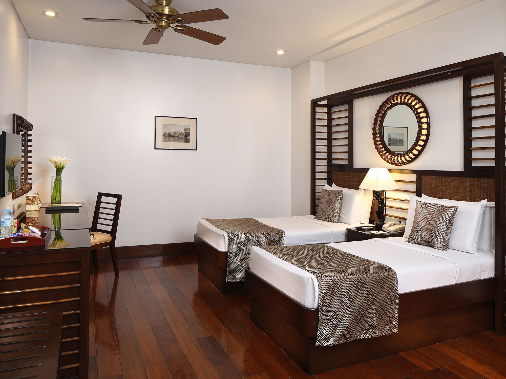 Ein Hotelzimmer mit zwei Einzelbetten, einem Schreibtisch mit Stuhl, einem Deckenventilator, einem gerahmten Bild an der Wand und einem Nachttisch mit Lampe.