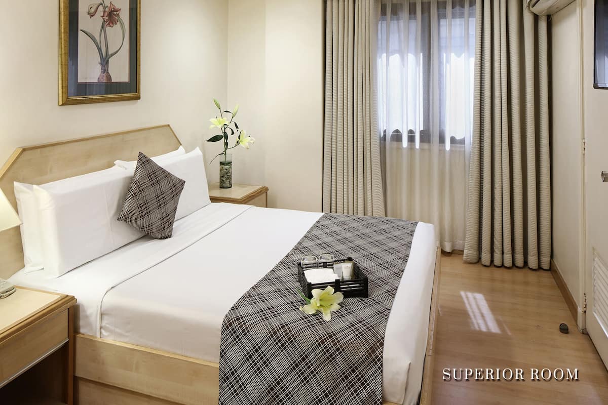 A hotel room with a bed, a nightstand, a bedside table, a framed picture on the wall, a window with curtains, and an air conditioning unit.