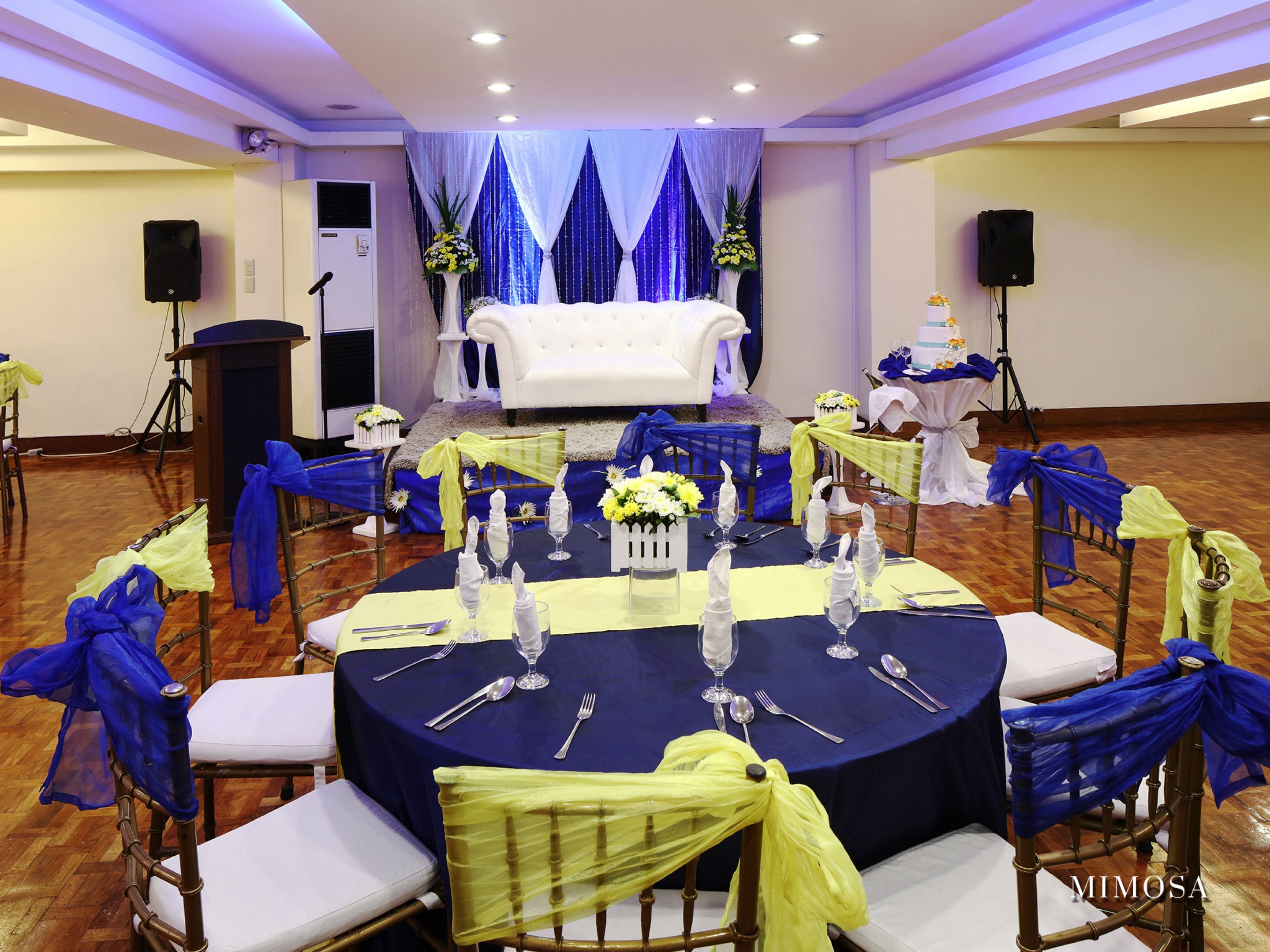 A banquet hall with round tables set for a formal event, featuring white chairs with blue and yellow sashes, and a white sofa in the background.