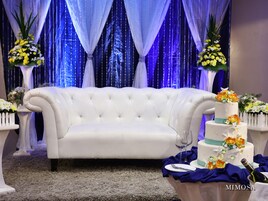 Um sofá branco com detalhes em tufos numa sala com decoração azul e branca, um bolo de três andares com decorações florais, e uma garrafa de champanhe sobre uma mesa.