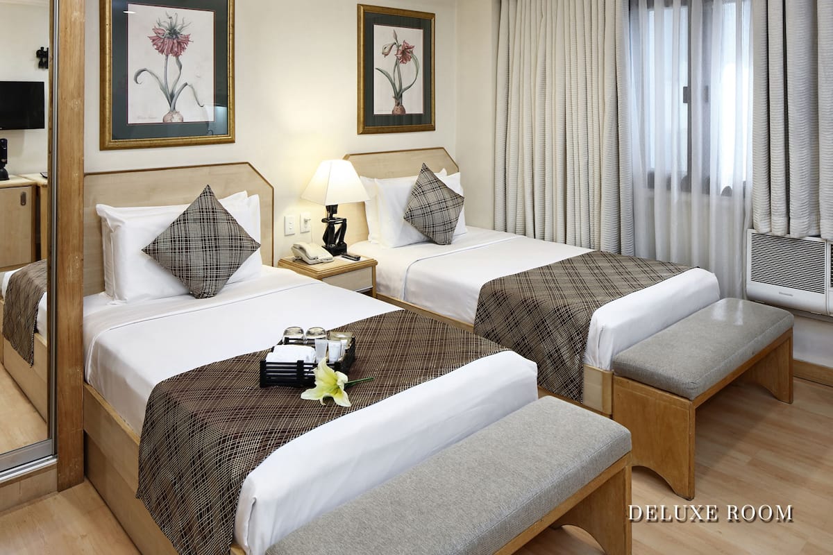 A DELUXE ROOM with two beds, a bedside table with a lamp, a mirror, and a window with curtains.