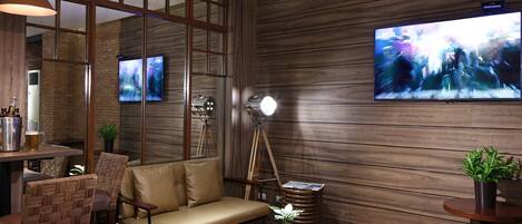 A modern lounge area with a sofa, chairs, a small table with a plant, a bar area with bottles, and two mounted televisions.