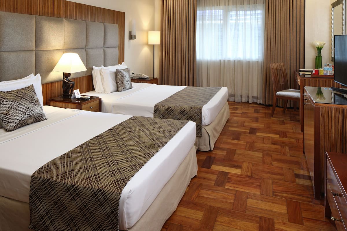 A hotel room with two beds, a desk with a TV, a chair, and a window with curtains.