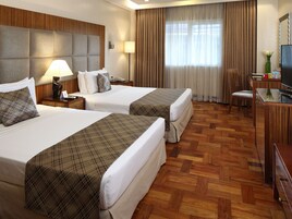 Quarto de hotel com duas camas, uma escrivaninha com televisão, uma cadeira e uma janela com cortinas.